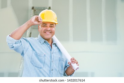 Hispanic Male Contractor With Blueprint Plans Wearing Hard Hat In Front Of Drywall And Ladder.