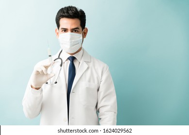 Hispanic Male Doctor Holding Syringe While Wearing Mask Against Isolated Background