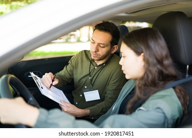 Hispanic Male Instructor Giving Instructions To A Teenage Girl Before Starting Her Driving Practice. Young Man Preparing To Grade An Adolescent Girl