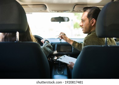 Hispanic Male Instructor And Teenage Girl Seen From Behind Inside A Car While Having A Driver License Exam. Caucasian Adolescent Girl Doing A Driving Test