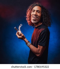 Hispanic Male Smoking Marijuana From A Bong On A Dark Background Illuminated By Blue Light, Get High. Cannabis Legalization Concept