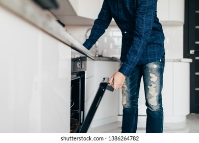 Hispanic Male Wearing Blue Shirt In Modern Kitchen Leaning Towards Open Oven Door Holding A Fork Looking Past Camera.