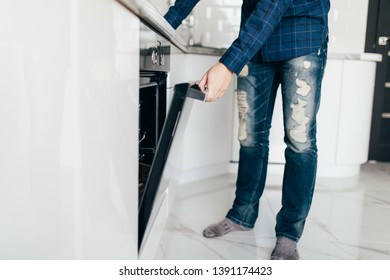 Hispanic Male Wearing Blue Shirt In Modern Kitchen Leaning Towards Open Oven Door Holding A Fork Looking Past Camera.