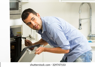 Hispanic Male Wearing Blue Shirt In Modern Kitchen Leaning Towards Open Oven Door Holding A Fork Looking Past Camera.