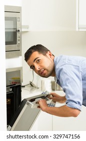 Hispanic Male Wearing Blue Shirt In Modern Kitchen Leaning Towards Open Oven Door Holding A Fork Looking Past Camera.