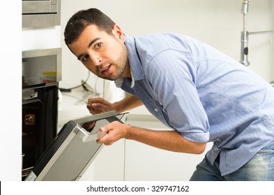 Hispanic Male Wearing Blue Shirt In Modern Kitchen Leaning Towards Open Oven Door Holding A Fork Looking Past Camera.