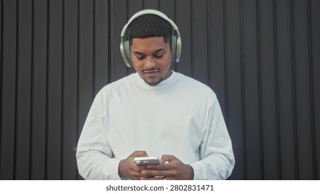 Hispanic man with beard adjusts overear headphones and holds smartphone against dark metal building wall; focused listening.