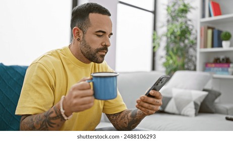 Hispanic man with beard drinking coffee and using smartphone in a modern living room.