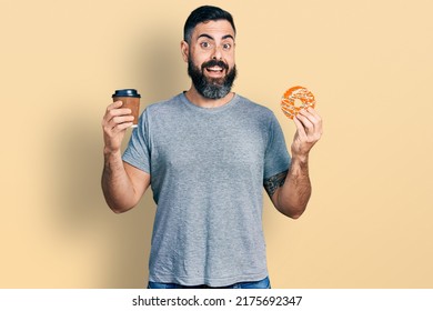 Hispanic man with beard eating doughnut and drinking coffee celebrating crazy and amazed for success with open eyes screaming excited.  - Powered by Shutterstock