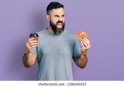 Hispanic man with beard eating doughnut and drinking coffee angry and mad screaming frustrated and furious, shouting with anger. rage and aggressive concept.  - Shutterstock ID 2186245553