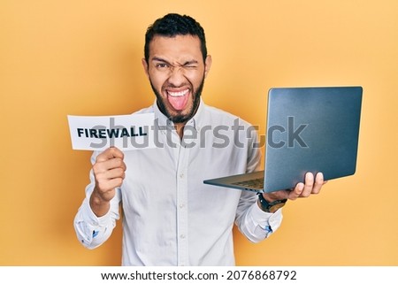 Hispanic man with beard holding computer laptop and firewall banner sticking tongue out happy with funny expression. 