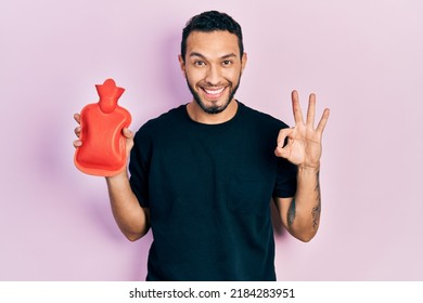 Hispanic man with beard holding hot water bag doing ok sign with fingers, smiling friendly gesturing excellent symbol 