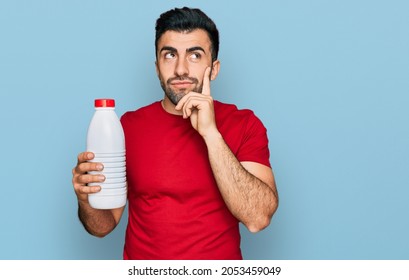 Hispanic Man With Beard Holding Liter Bottle Of Milk Serious Face Thinking About Question With Hand On Chin, Thoughtful About Confusing Idea 