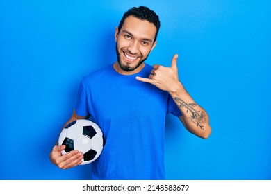 Hispanic man with beard holding soccer ball smiling doing phone gesture with hand and fingers like talking on the telephone. communicating concepts. 