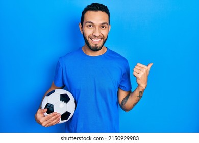 Hispanic man with beard holding soccer ball smiling with happy face looking and pointing to the side with thumb up. 