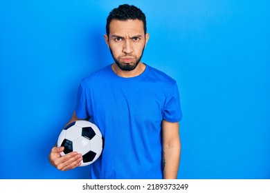 Hispanic Man With Beard Holding Soccer Ball Puffing Cheeks With Funny Face. Mouth Inflated With Air, Crazy Expression. 