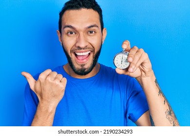 Hispanic man with beard holding stopwatch pointing thumb up to the side smiling happy with open mouth 