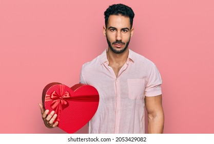 Hispanic Man With Beard Holding Valentine Gift Thinking Attitude And Sober Expression Looking Self Confident 