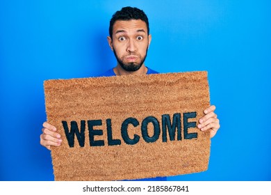 Hispanic Man With Beard Holding Welcome Doormat Puffing Cheeks With Funny Face. Mouth Inflated With Air, Catching Air. 