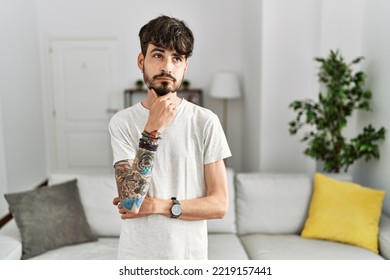 Hispanic Man With Beard At The Living Room At Home With Hand On Chin Thinking About Question, Pensive Expression. Smiling With Thoughtful Face. Doubt Concept. 