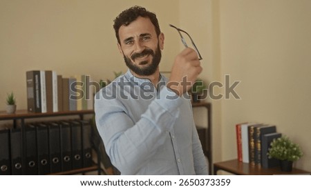 Hispanic man with a beard in an office setting removes glasses and smiles near shelves with books and plants.