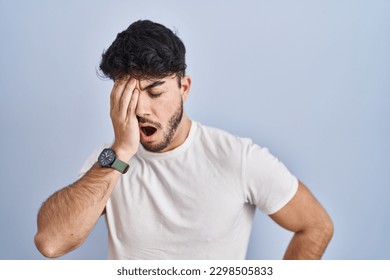 Hispanic man with beard standing over white background yawning tired covering half face, eye and mouth with hand. face hurts in pain. 