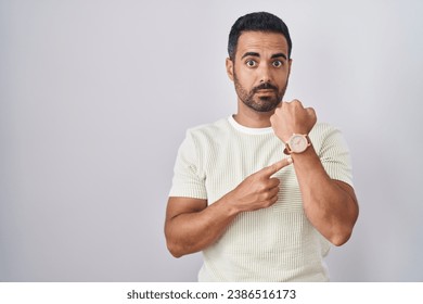 Hispanic man with beard standing over isolated background in hurry pointing to watch time, impatience, looking at the camera with relaxed expression 