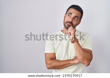 Hispanic man with beard standing over isolated background with hand on chin thinking about question, pensive expression. smiling with thoughtful face. doubt concept. 