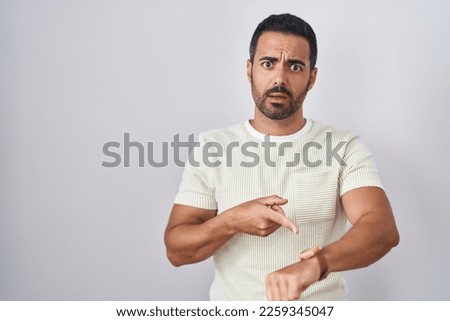 Hispanic man with beard standing over isolated background in hurry pointing to watch time, impatience, upset and angry for deadline delay 