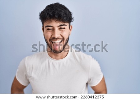 Hispanic man with beard standing over white background sticking tongue out happy with funny expression. emotion concept. 