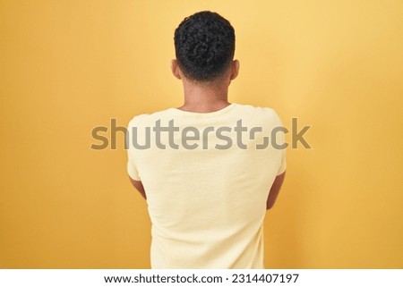 Hispanic man with beard standing over yellow background standing backwards looking away with crossed arms 