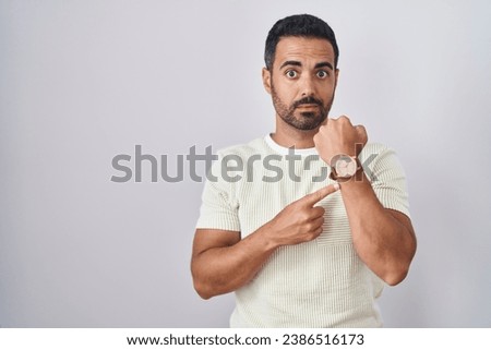 Hispanic man with beard standing over isolated background in hurry pointing to watch time, impatience, looking at the camera with relaxed expression 