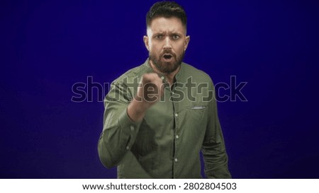 Hispanic man with beard thrusts clenched fist toward camera in studio with olive shirt, stern gaze and scowl; anger confrontation.
