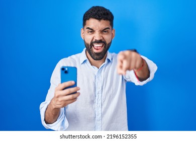 Hispanic Man With Beard Using Smartphone Typing Message Pointing Displeased And Frustrated To The Camera, Angry And Furious With You 