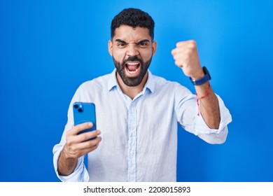 Hispanic Man With Beard Using Smartphone Typing Message Angry And Mad Raising Fist Frustrated And Furious While Shouting With Anger. Rage And Aggressive Concept. 