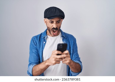 Hispanic man with beard using smartphone typing message angry and mad screaming frustrated and furious, shouting with anger. rage and aggressive concept.  - Shutterstock ID 2363789381
