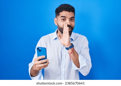 Hispanic man with beard using smartphone typing message hand on mouth telling secret rumor, whispering malicious talk conversation 