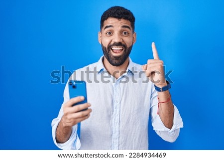 Hispanic man with beard using smartphone typing message pointing finger up with successful idea. exited and happy. number one. 