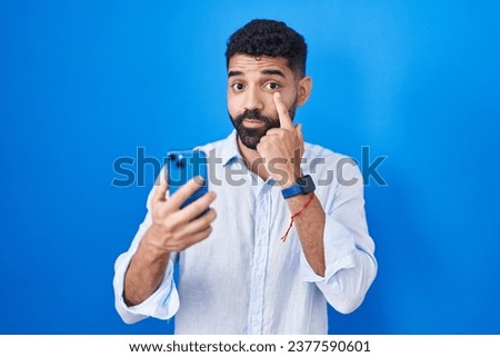 Hispanic man with beard using smartphone typing message pointing to the eye watching you gesture, suspicious expression 