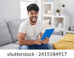 Hispanic man with beard using touchpad sitting on the sofa sticking tongue out happy with funny expression. emotion concept. 