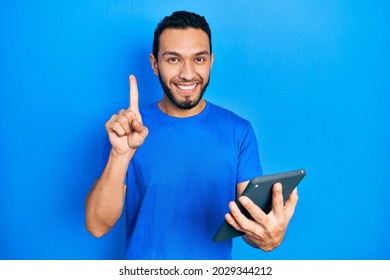 Hispanic man with beard using touchpad device smiling with an idea or question pointing finger with happy face, number one 