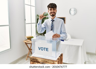 Hispanic Man With Beard Voting Putting Envelop In Ballot Box Smiling And Confident Gesturing With Hand Doing Small Size Sign With Fingers Looking And The Camera. Measure Concept. 