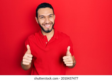 Hispanic Man With Beard Wearing Casual Red T Shirt Success Sign Doing Positive Gesture With Hand, Thumbs Up Smiling And Happy. Cheerful Expression And Winner Gesture. 