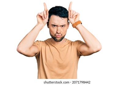 Hispanic Man With Beard Wearing Casual T Shirt Doing Funny Gesture With Finger Over Head As Bull Horns 