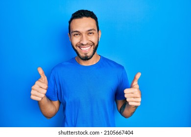 Hispanic man with beard wearing casual blue t shirt success sign doing positive gesture with hand, thumbs up smiling and happy. cheerful expression and winner gesture. 