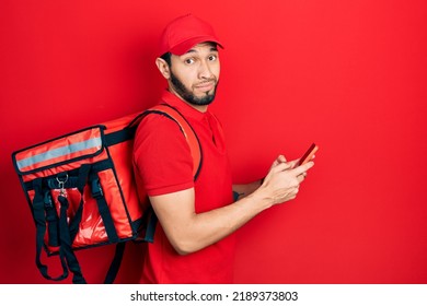 Hispanic Man With Beard Wearing Delivery Package Backpack Using Smartphone Clueless And Confused Expression. Doubt Concept. 