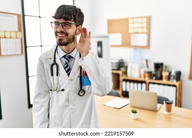 Hispanic Man With Beard Wearing Doctor Uniform And Stethoscope At The Office Smiling With Hand Over Ear Listening An Hearing To Rumor Or Gossip. Deafness Concept. 