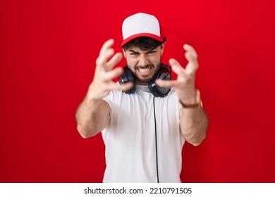 Hispanic Man With Beard Wearing Gamer Hat And Headphones Shouting Frustrated With Rage, Hands Trying To Strangle,