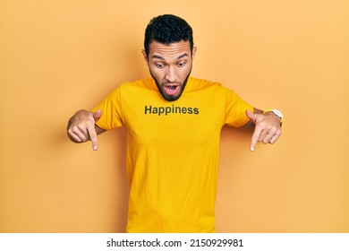 Hispanic Man With Beard Wearing T Shirt With Happiness Word Message Pointing Down With Fingers Showing Advertisement, Surprised Face And Open Mouth 