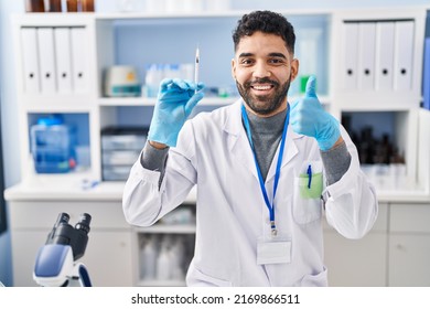 Hispanic Man With Beard Working At Scientist Laboratory Holding Syringe Smiling Happy And Positive, Thumb Up Doing Excellent And Approval Sign 